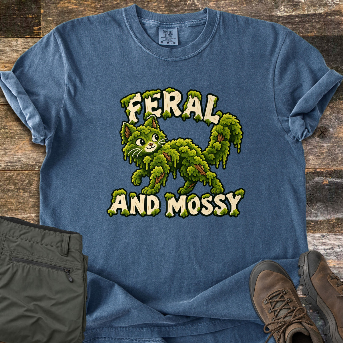 Feral and Mossy Comfort Colors T-shirt in Denim, laid flat. A green cartoon cat made of dripping moss walks between the words Feral and Mossy in cream lettering. Sleeves rolled, on a wood plank floor beside olive cargo pants and brown hiking boots.