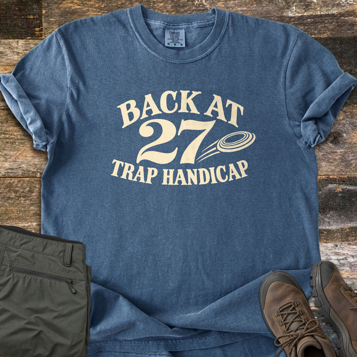 Back at 27 Trap Handicap Comfort Colors T-shirt in Denim, laid flat. Cream text reads Back at 27 above Trap Handicap, with a flying clay target beside the 27. The shirt lies on weathered wood planks with folded olive pants and brown hiking boots.