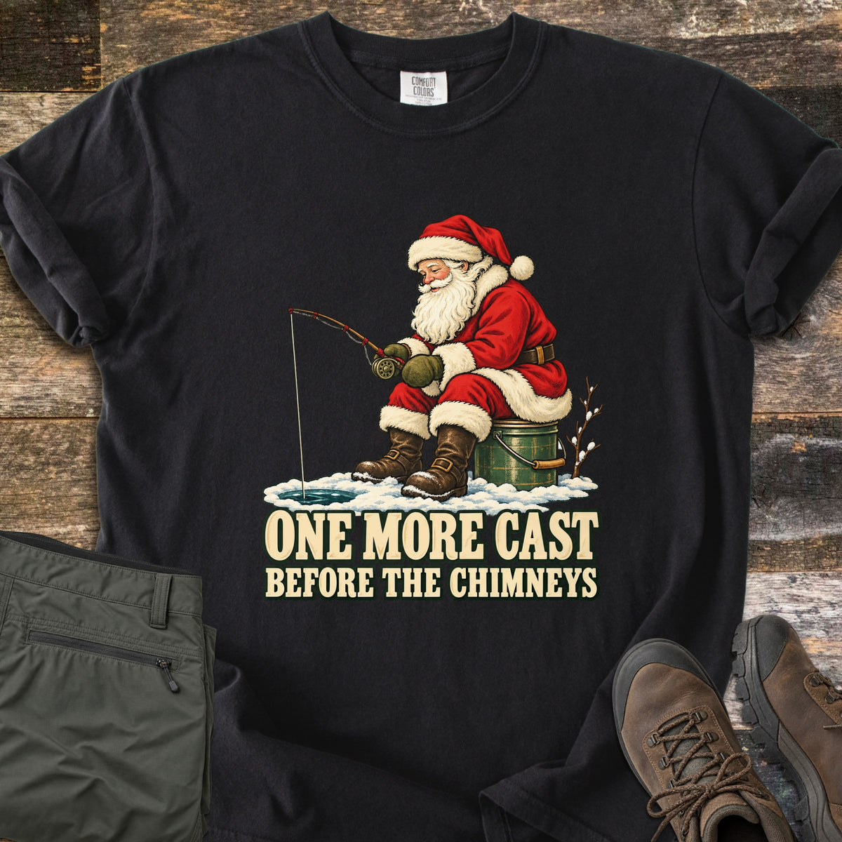 One More Cast Before the Chimneys Comfort Colors T-shirt in Black, laid flat. Santa in his red suit sits on a green bucket, fishing through a hole in the ice, above the words One More Cast Before the Chimneys. Folded olive pants and brown hiking boots rest on weathered wood.