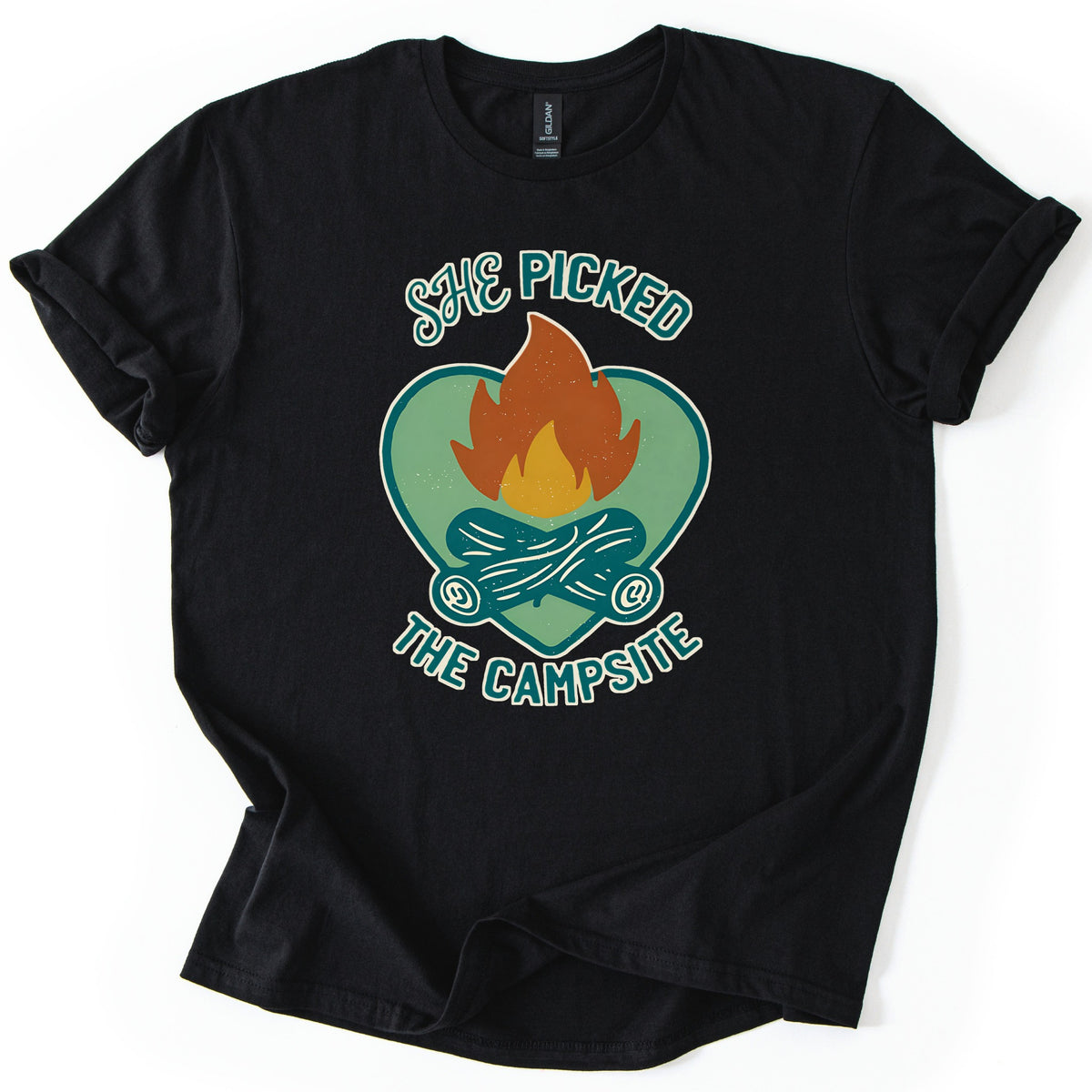 She Picked The Campsite Comfort Colors T-shirt in Black, laid flat. An orange and yellow campfire on crossed teal logs inside a sage green heart, with the words SHE PICKED above and THE CAMPSITE below. The shirt lies on a white surface with its sleeves cuffed.