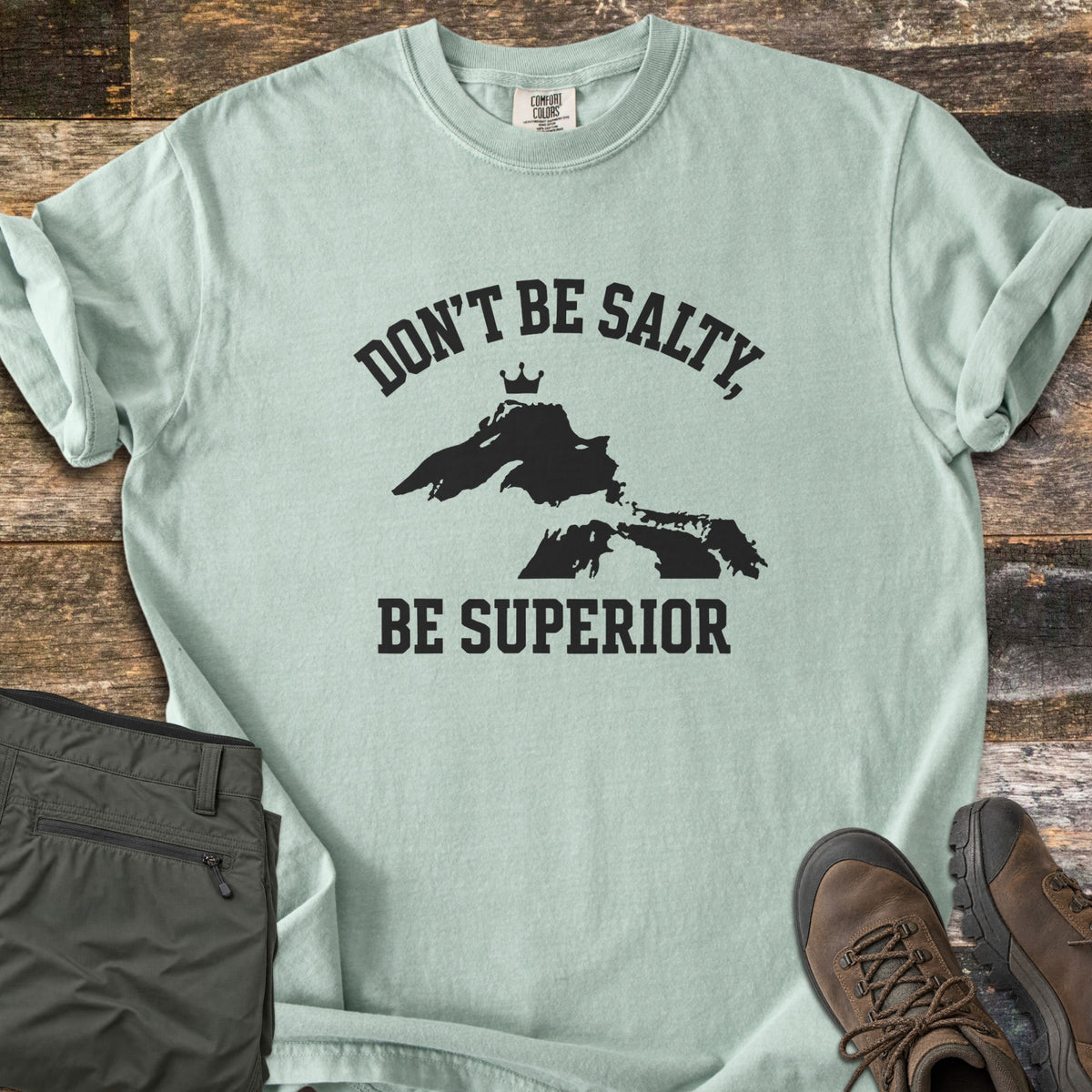 Be Superior Comfort Colors T-shirt in Bay, laid flat. Black text reads Don't Be Salty, arched over a Lake Superior silhouette topped with a small crown, with Be Superior below. Sleeves rolled, on weathered wood with olive hiking pants and brown hiking boots.