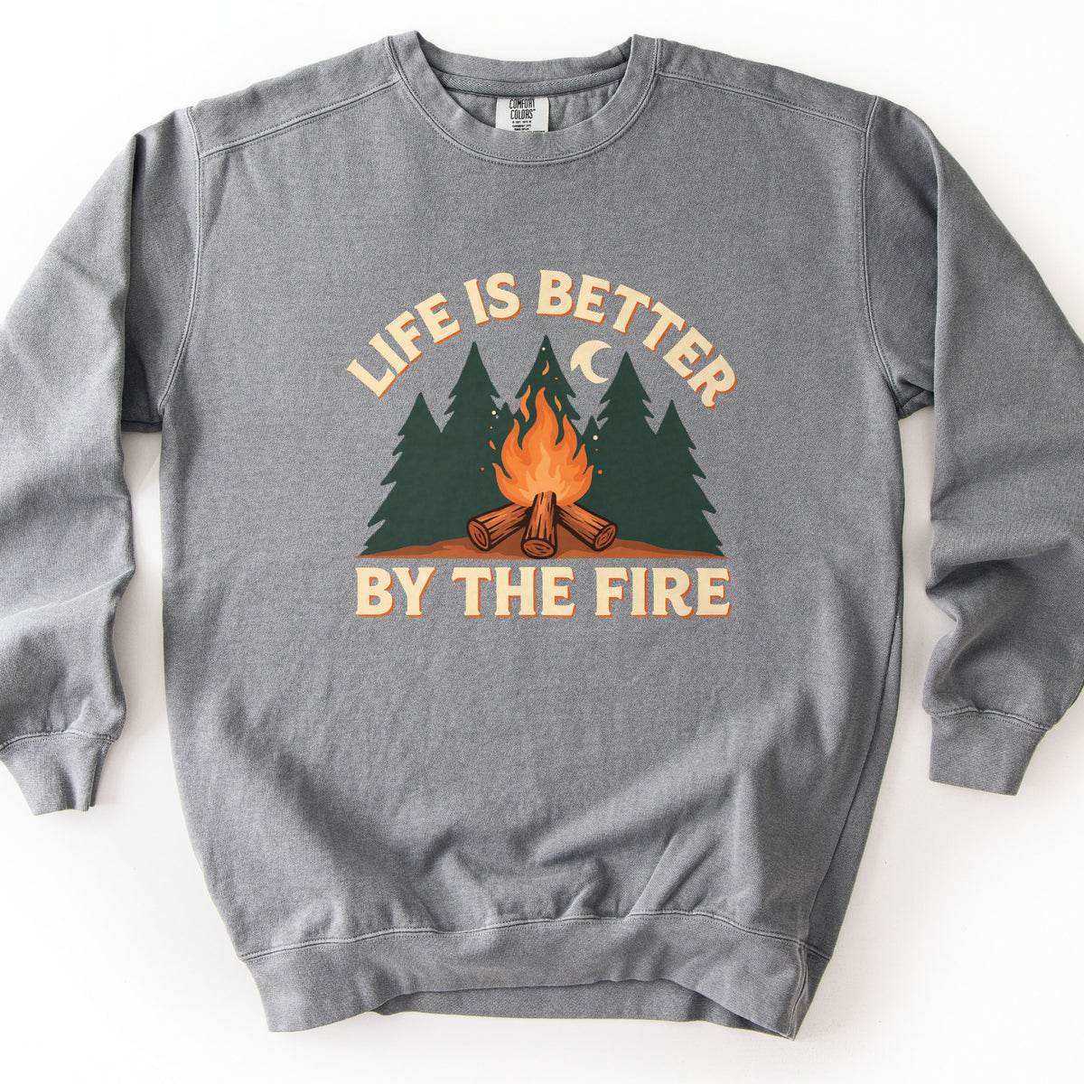 Life Is Better By The Fire Comfort Colors crewneck sweatshirt in Grey, laid flat. Cream letters with an orange shadow arch Life Is Better over dark green pine trees, a crescent moon and a campfire of crossed logs, with By The Fire below. It lies on a white surface.