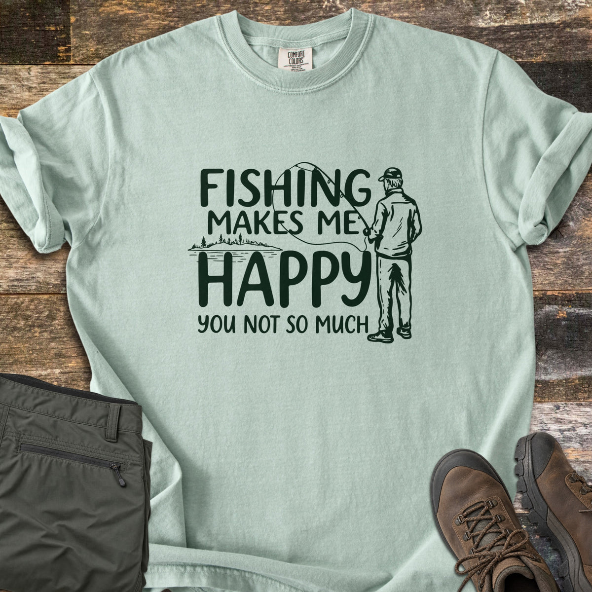 Fishing Makes Me Happy Comfort Colors T-shirt in Bay, laid flat. Dark green print of a man in a cap casting a fishing rod by a tree-lined lake, with the words Fishing Makes Me Happy You Not So Much. Laid on weathered wood with olive pants and brown hiking boots.