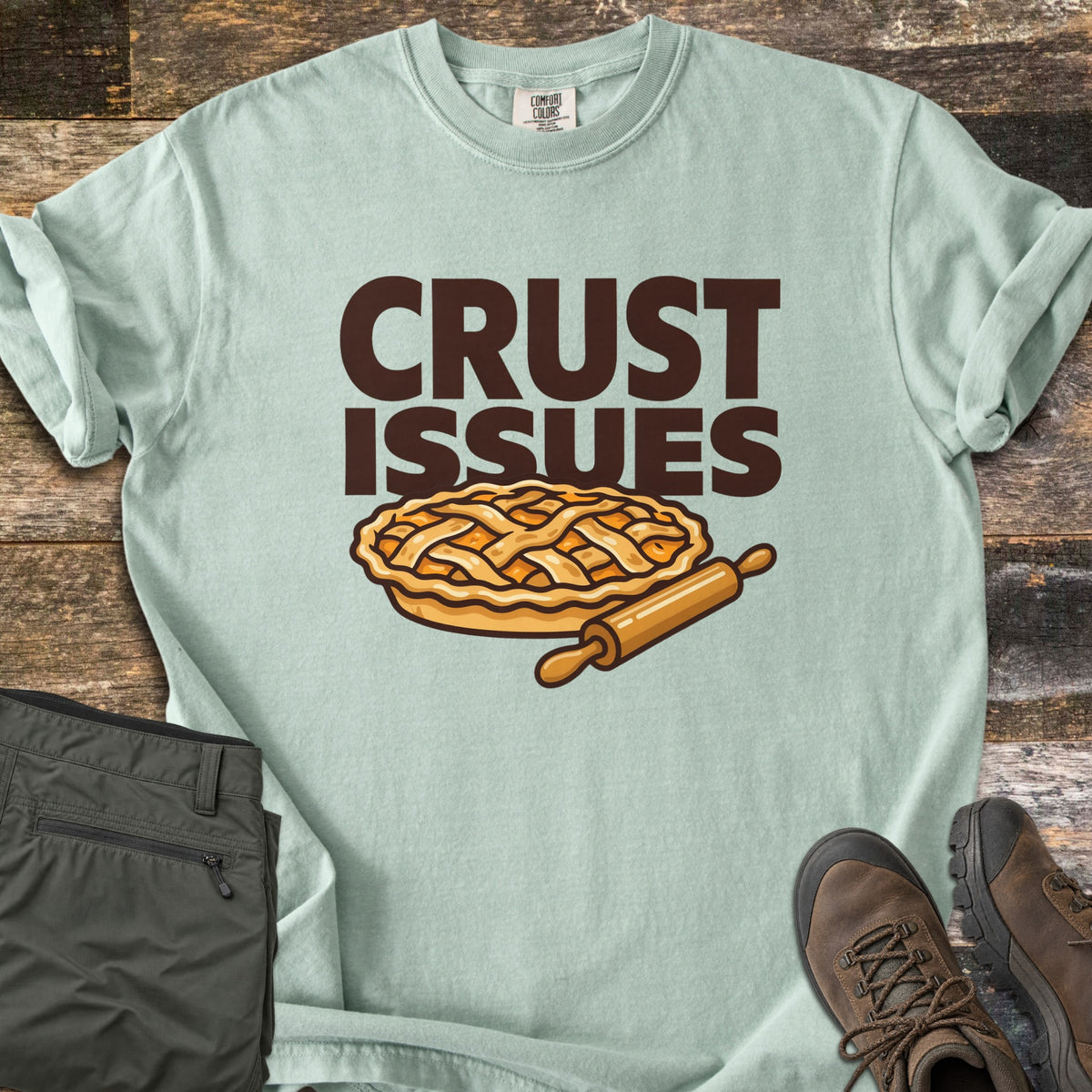 Crust Issues Comfort Colors T-shirt in Bay, laid flat. The words CRUST ISSUES in dark brown block letters sit above a cartoon lattice-top pie and a wooden rolling pin. Olive pants and brown hiking boots lie beside it on a rustic wood surface.