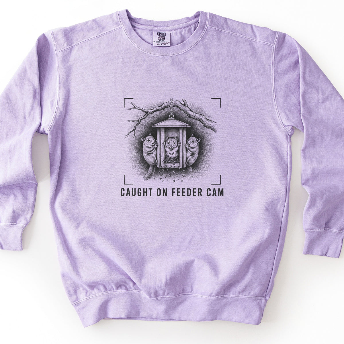 Caught On Feeder Cam Comfort Colors crewneck sweatshirt in Orchid, laid flat. A black sketch of three squirrels holding onto a wooden bird feeder hanging from a branch, with seeds falling and camera viewfinder corners around it, above the words Caught On Feeder Cam. Shown on a white surface.