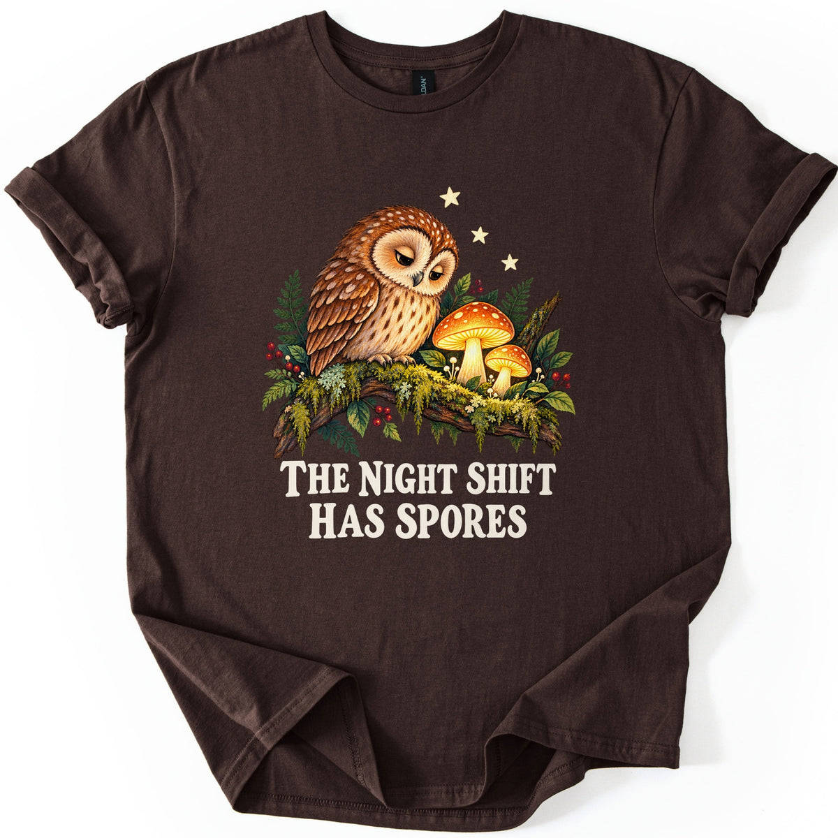 The Night Shift Has Spores Comfort Colors T-shirt in Dark Chocolate, laid flat. A sleepy brown owl perches on a mossy branch beside two glowing orange mushrooms, with ferns, red berries and small stars, above the words The Night Shift Has Spores in cream. Shown on white.