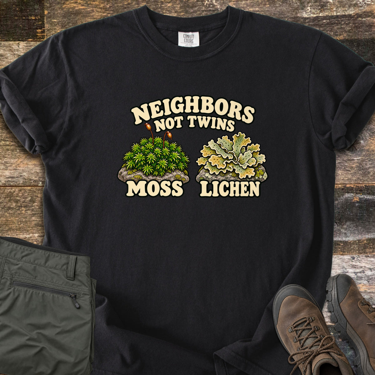 Neighbors Not Twins Comfort Colors T-shirt in Black, laid flat. Arched text Neighbors Not Twins over two illustrated rock clumps: green moss with brown stalks labeled Moss, and pale green leafy lichen labeled Lichen. Olive hiking pants and brown boots sit beside it on weathered wood.
