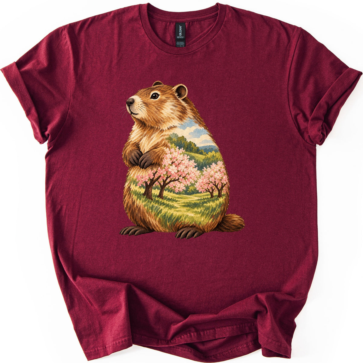 Groundhog Blossom Meadow Comfort Colors T-shirt in Maroon, laid flat. A painted groundhog sits upright with paws at its chest, its body filled with a meadow of pink blossoming trees, green hills and blue sky. No words. Sleeves cuffed, on a white background.