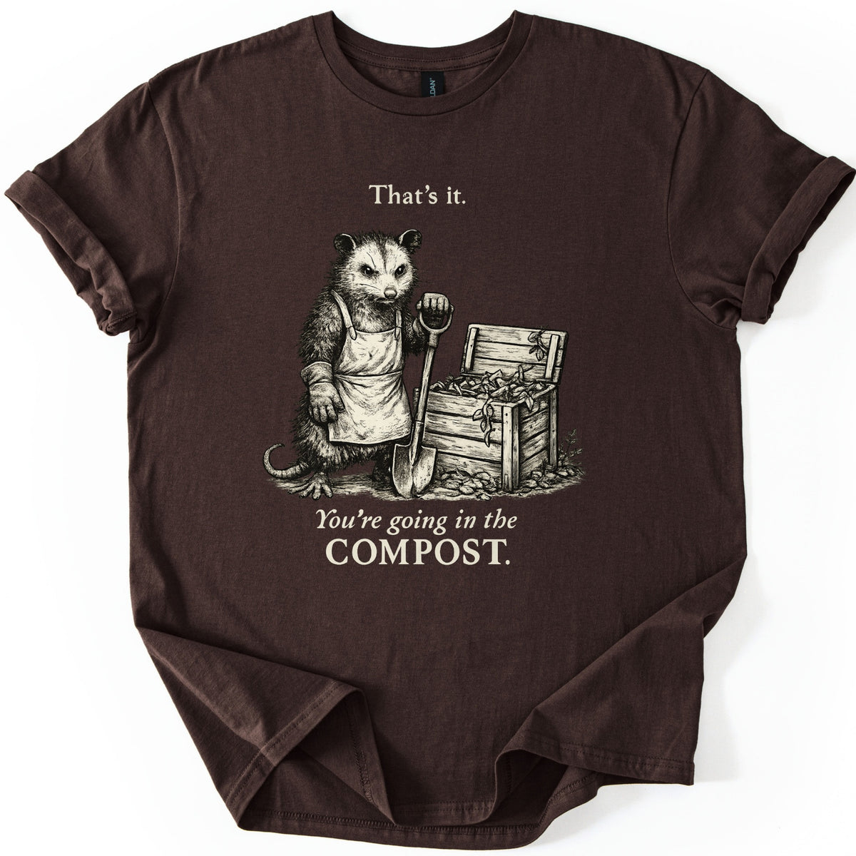Going In The Compost Possum Comfort Colors T-shirt in Dark Chocolate, laid flat. A scowling possum in an apron and work glove leans on a shovel beside an open wooden compost bin, between the words That's it. and You're going in the COMPOST. Cream print, shirt on white with cuffed sleeves.