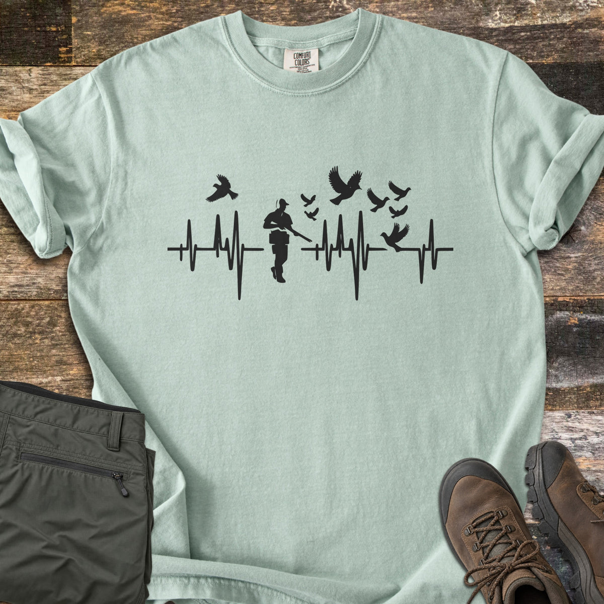 Bird Hunter Comfort Colors T-shirt in Bay, laid flat. A black heartbeat line crosses the chest with a hunter silhouette in a cap holding a shotgun and birds flying up from the line. Folded olive pants and brown hiking boots lie beside it on weathered wood.