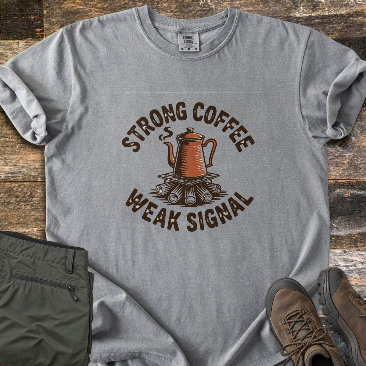 Strong Coffee Weak Signal Comfort Colors T-shirt in Grey, laid flat. A steaming copper coffee pot sits on a grate over campfire logs, arched by the words Strong Coffee above and Weak Signal below. Folded olive hiking pants and brown hiking boots lie on weathered wood planks.