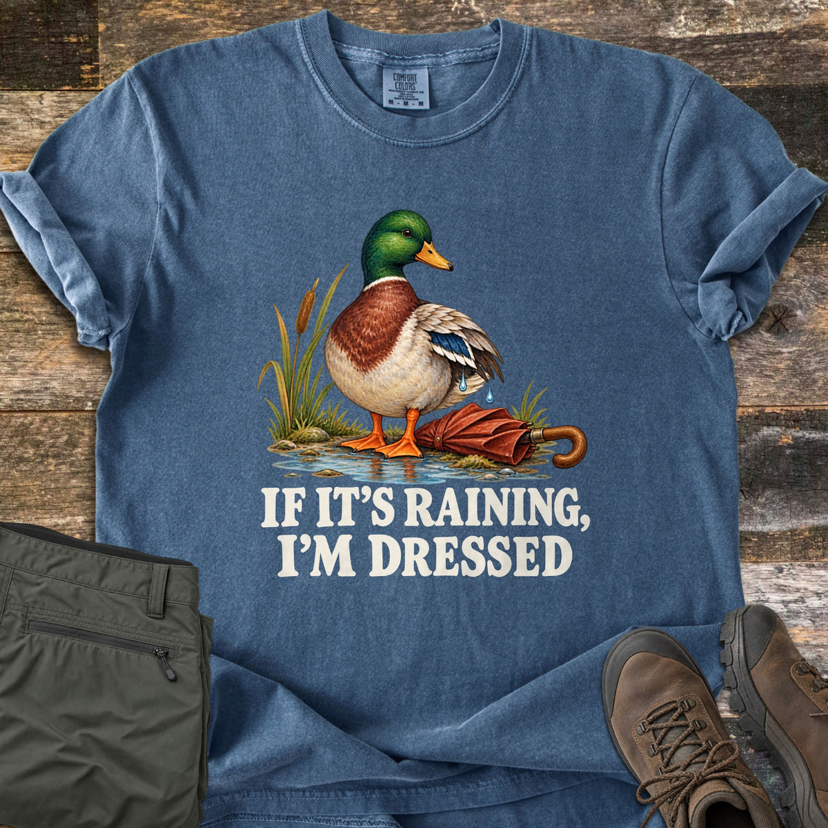 If It's Raining, I'm Dressed Comfort Colors T-shirt in Denim, laid flat. A mallard duck with water dripping off its wings stands in a puddle by cattails next to a closed red umbrella, above the words If It's Raining, I'm Dressed. On weathered wood with olive pants and brown hiking boots.
