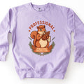 Professional Pinecone Inspector Comfort Colors crewneck sweatshirt in Orchid, laid flat. A cartoon squirrel in an orange cap studies a pinecone through a magnifying glass under the arched rust word Professional, above a banner reading Pinecone Inspector framed by pine branches. On a white surface.