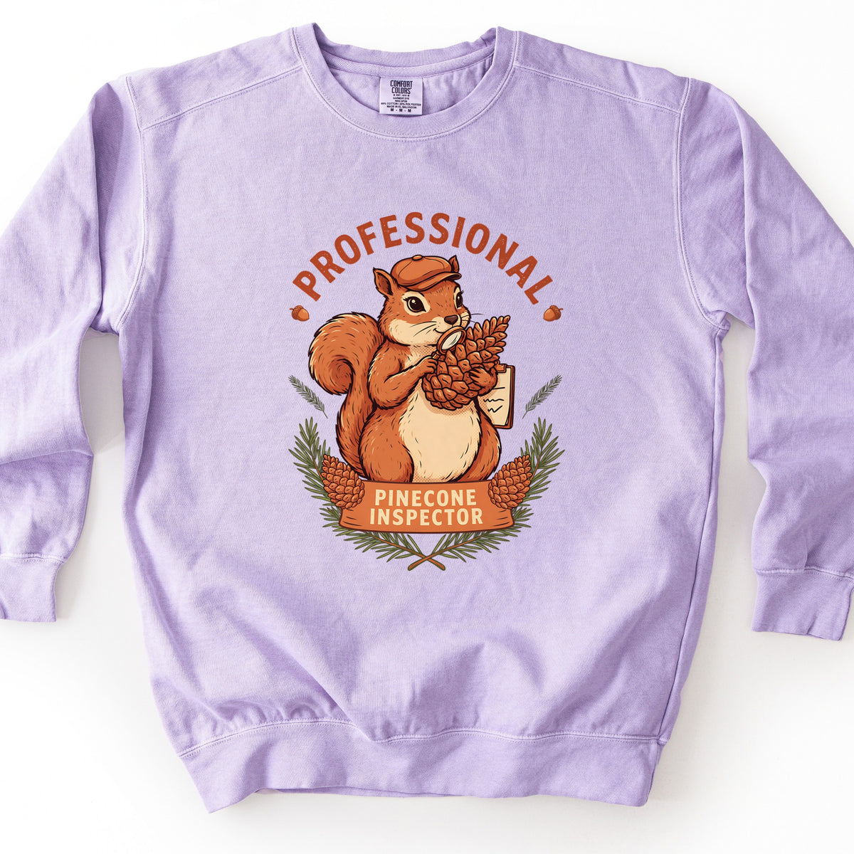 Professional Pinecone Inspector Comfort Colors crewneck sweatshirt in Orchid, laid flat. A cartoon squirrel in an orange cap studies a pinecone through a magnifying glass under the arched rust word Professional, above a banner reading Pinecone Inspector framed by pine branches. On a white surface.