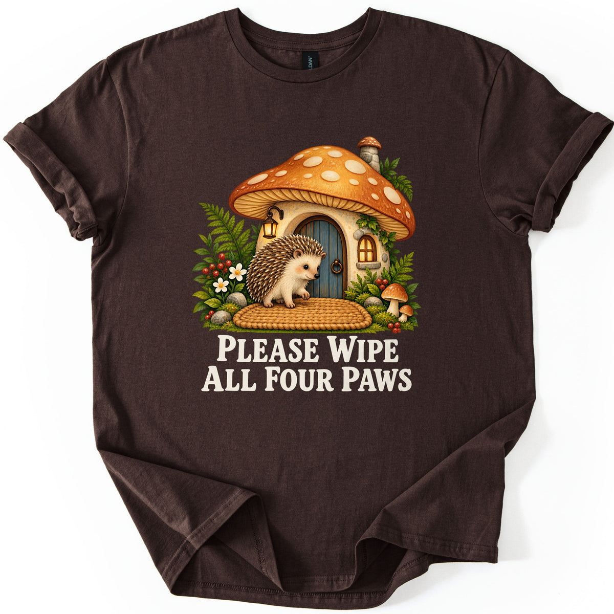 Please Wipe All Four Paws Comfort Colors T-shirt in Dark Chocolate, laid flat. A hedgehog stands on a woven doormat outside a mushroom cottage with an orange spotted cap, lantern and blue door, surrounded by ferns and small mushrooms. Below, cream text reads Please Wipe All Four Paws.