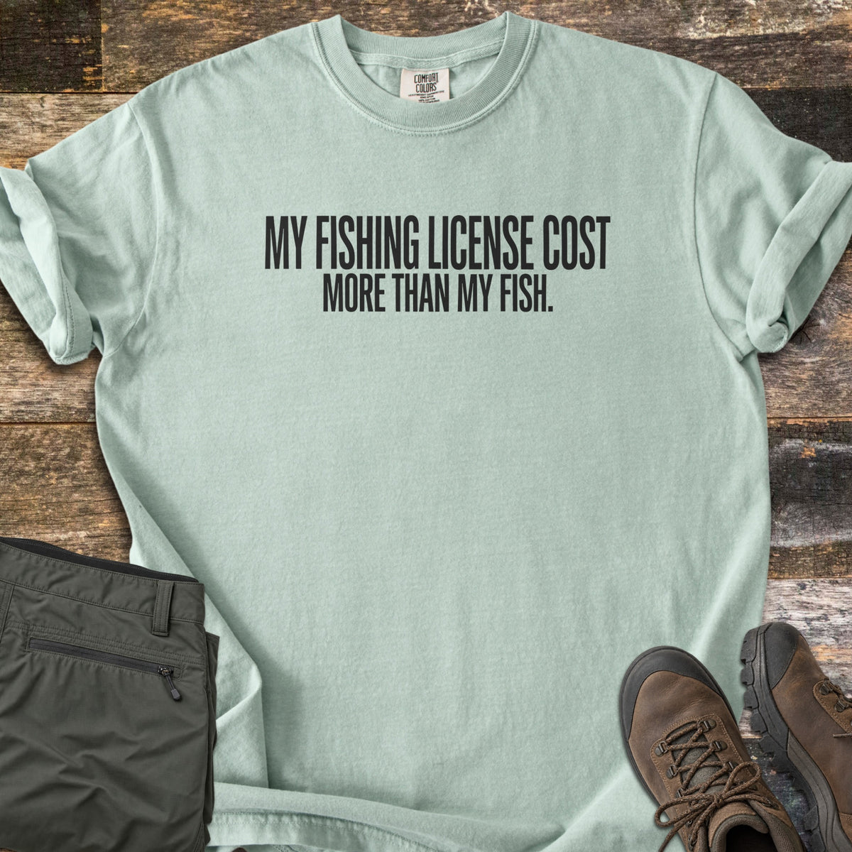 License Cost More Than My Fish Comfort Colors T-shirt in Bay, laid flat. Black block letters read My fishing license cost more than my fish. The shirt has rolled sleeves and lies on weathered wood planks beside folded olive hiking pants and brown hiking boots.