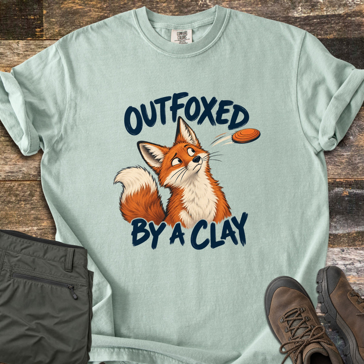 Outfoxed by a Clay Comfort Colors T-shirt in Bay, laid flat. A cartoon red fox glances sideways at an orange clay target flying past, between the words Outfoxed By A Clay in navy brush letters. Sleeves rolled on rustic wood with olive pants and brown hiking boots.