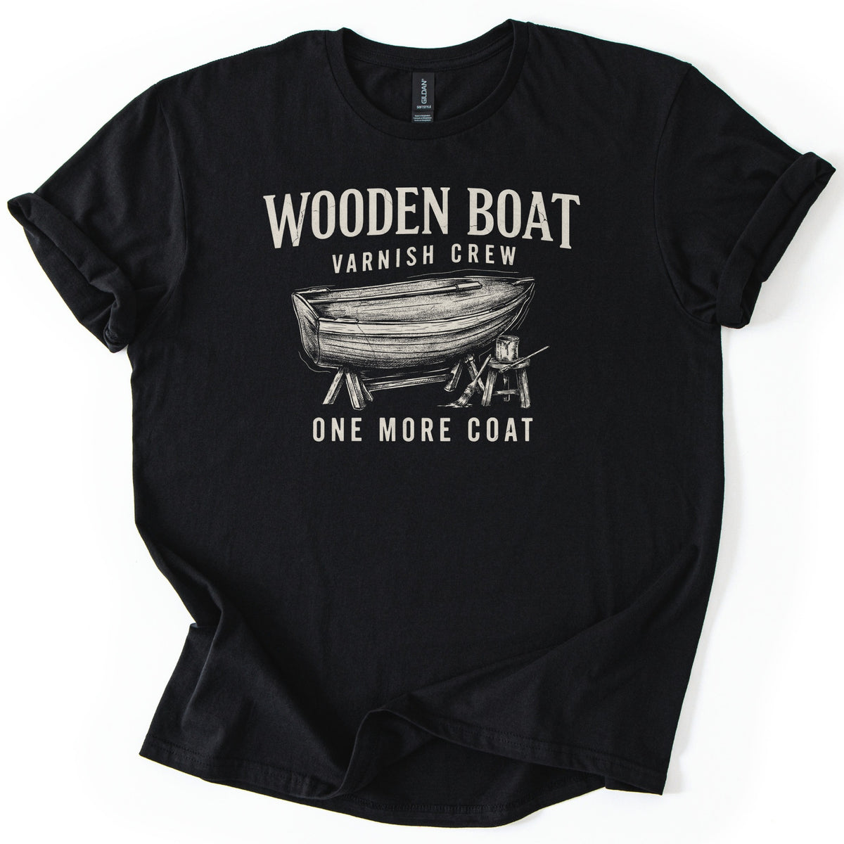 Wooden Boat Varnish Crew Comfort Colors T-shirt in Black, laid flat. Cream print reads Wooden Boat, Varnish Crew above a sketch of a wooden rowboat on sawhorses beside a stool with a varnish can and brush, then One More Coat. Sleeves cuffed on a white background.
