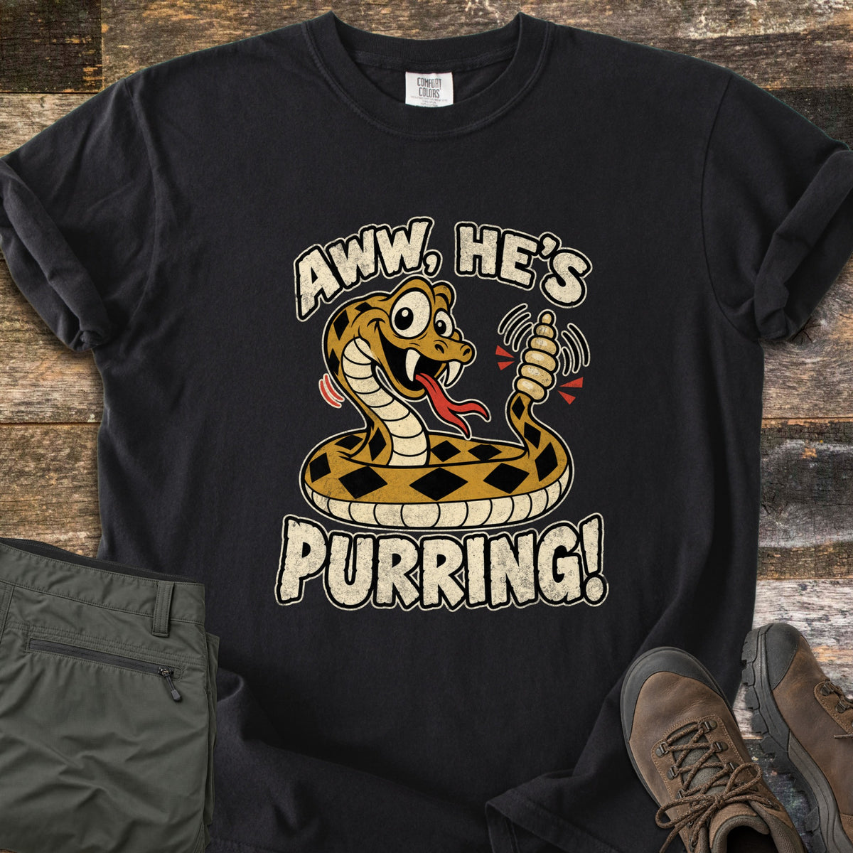 Aww He's Purring Comfort Colors T-shirt in Black, laid flat. A goofy, wide-eyed cartoon rattlesnake shakes its rattle between the cream-colored words Aww, He's Purring! Olive hiking pants and brown hiking boots lie beside the shirt on weathered wood boards.