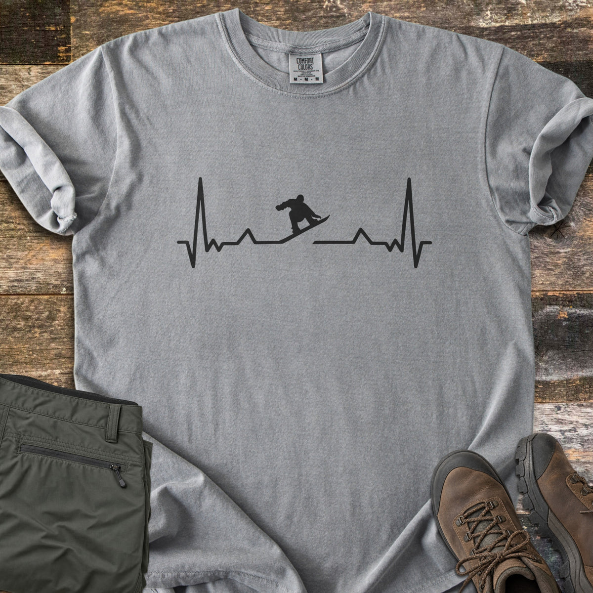 Snowboarder Heartbeat Comfort Colors T-shirt in Grey, laid flat. A black heartbeat line runs across the chest, with a snowboarder silhouette riding the line in the middle. Sleeves are rolled, on a rustic wood floor beside folded olive pants and brown hiking boots.
