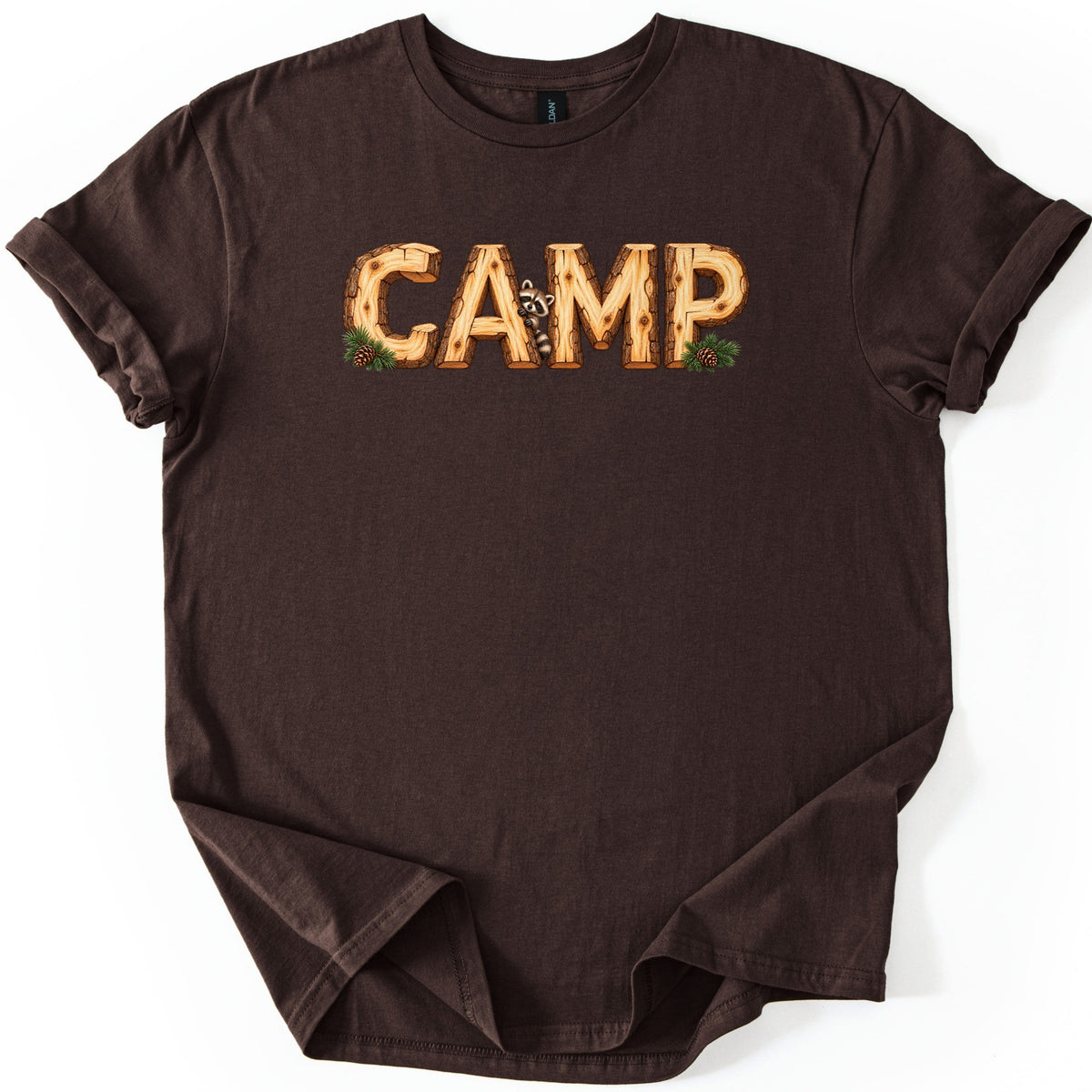 Camp Comfort Colors T-shirt in Dark Chocolate, laid flat. The word CAMP is printed in letters made of cut logs with bark and wood grain, a raccoon peeking out beside the A, and pine sprigs with pinecones at the ends. The shirt lies on white with rolled sleeves.