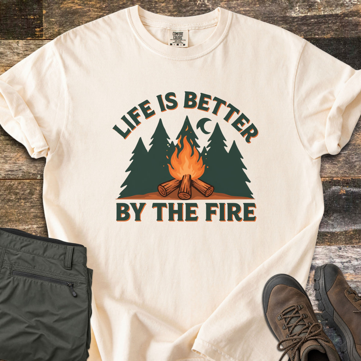 Life Is Better By The Fire Comfort Colors T-shirt in Ivory, laid flat. Arched dark green text reads Life Is Better over pine trees, a crescent moon and a campfire on crossed logs, with By The Fire below. Folded olive pants and brown hiking boots sit on weathered wood.