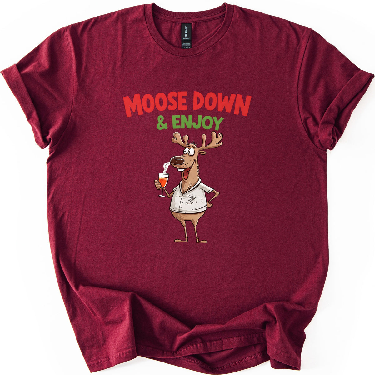 Moose Down And Enjoy Comfort Colors T-shirt in Maroon, laid flat. Red words Moose Down above green & Enjoy, over a grinning cartoon moose in a white short-sleeve shirt holding a glass of orange drink, hand on hip. Sleeves are cuffed on a white background.