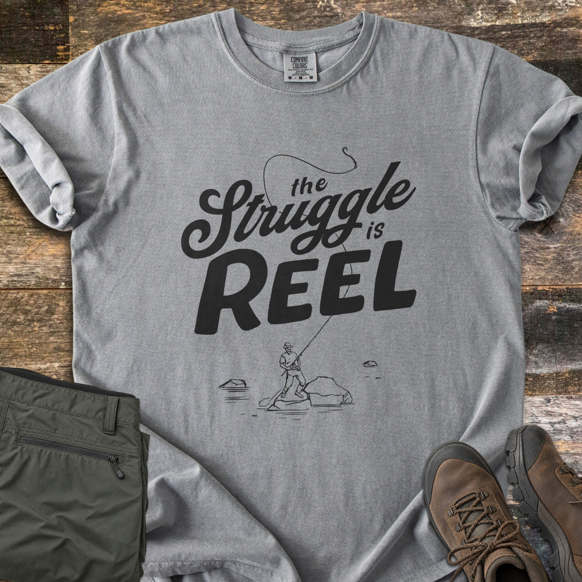 The Struggle Is Reel Comfort Colors T-shirt in Grey, laid flat. Black text reads the Struggle is REEL under an arcing fishing line with a hook, above a sketch of an angler casting from a rock in water. Folded olive pants and brown hiking boots sit on wood planks.