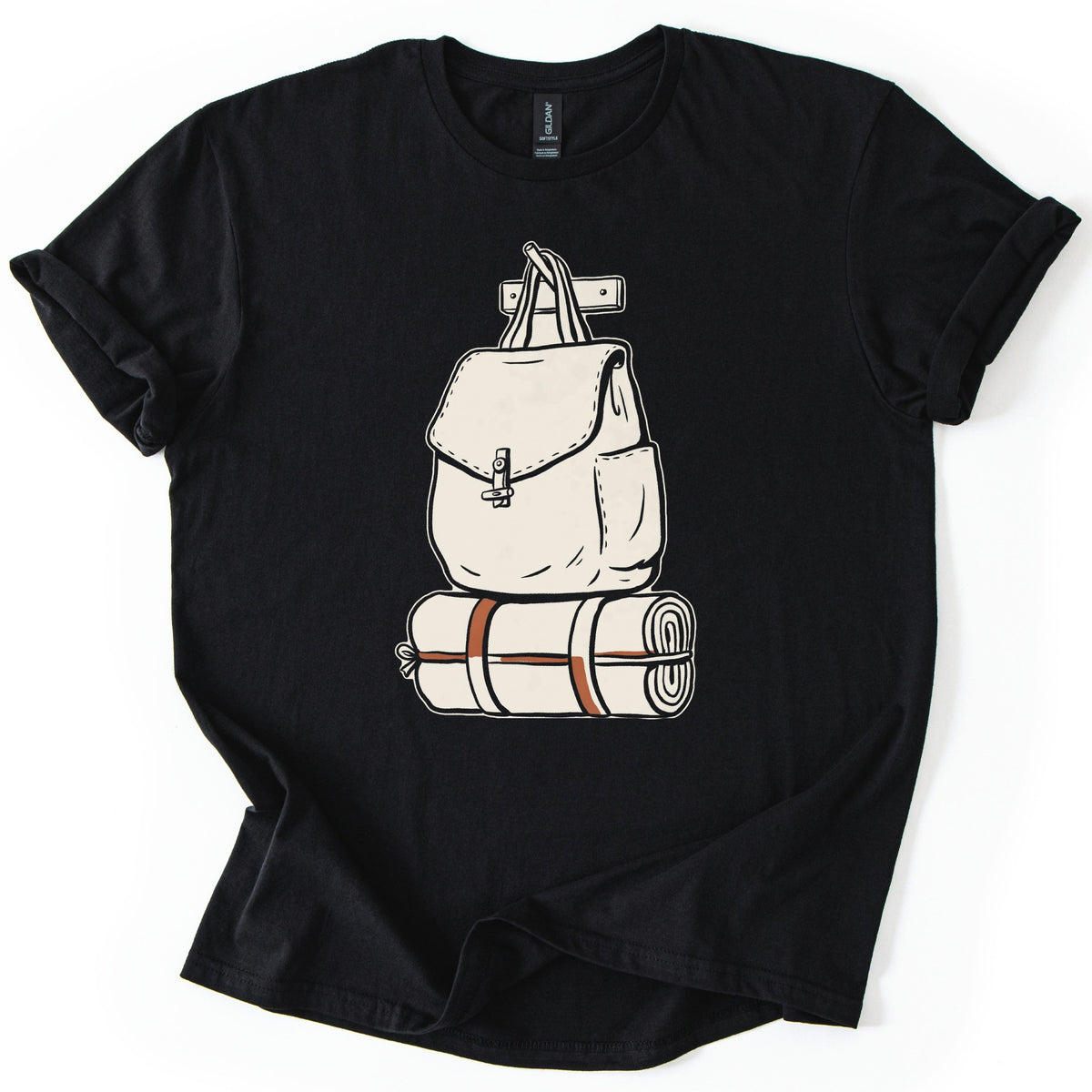 Backpack And Bedroll Comfort Colors T-shirt in Black, laid flat. A cream illustration of a canvas backpack with a toggle-clasp flap hangs by its handles from a wall peg above a rolled bedroll tied with rust-red straps. No words. Cuffed sleeves on a white surface.