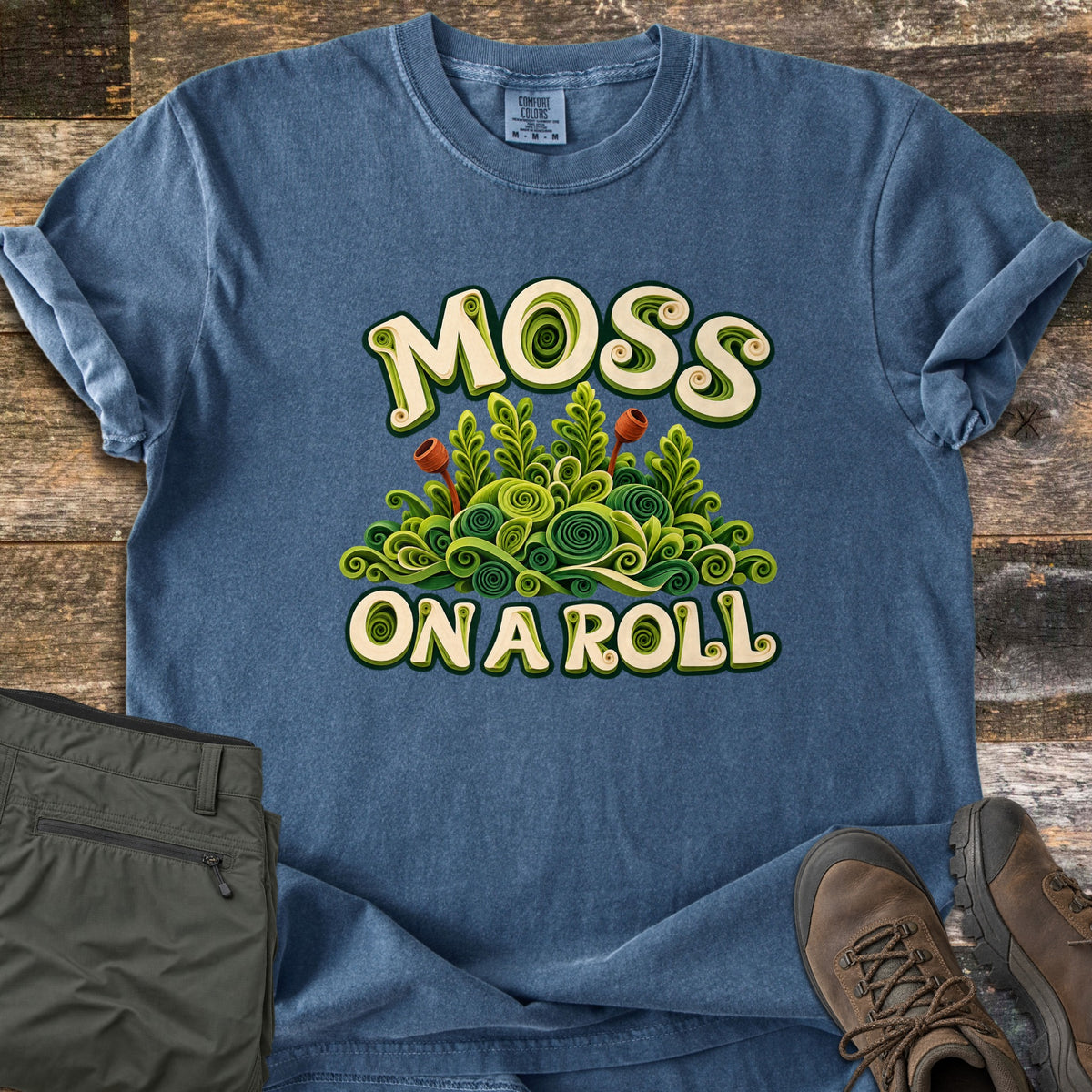 Moss on a Roll Comfort Colors T-shirt in Denim, laid flat. Quilled paper-style art of green moss with curled fronds and two rust-red spore stalks, between the words Moss and On a Roll. Olive cargo pants and brown hiking boots sit beside it on weathered wood planks.