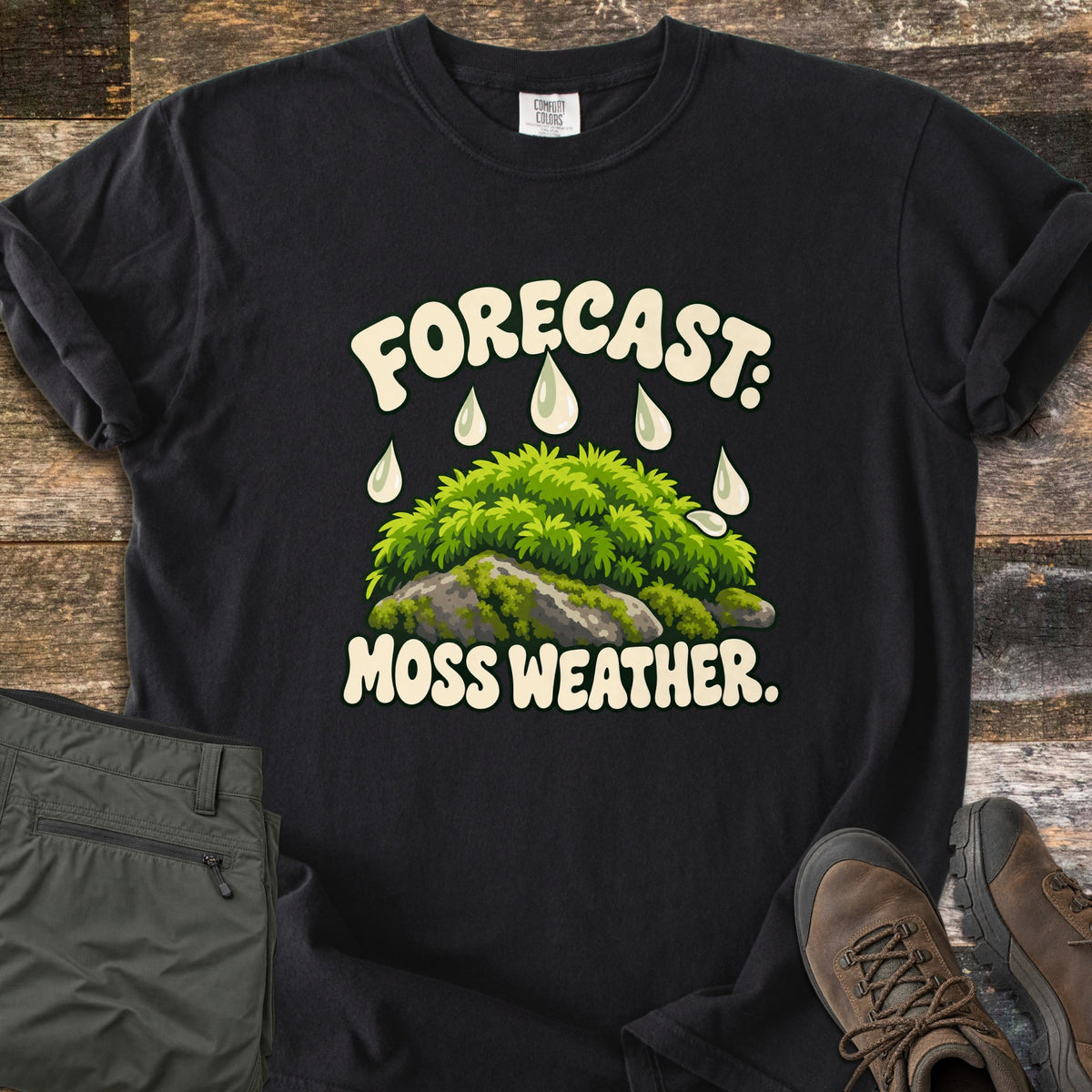 Forecast Moss Weather Comfort Colors T-shirt in Black, laid flat. A cartoon mound of bright green moss on a gray rock, with raindrops falling from the words Forecast: above and Moss Weather. below. Olive hiking pants and brown hiking boots sit beside it on weathered wood boards.