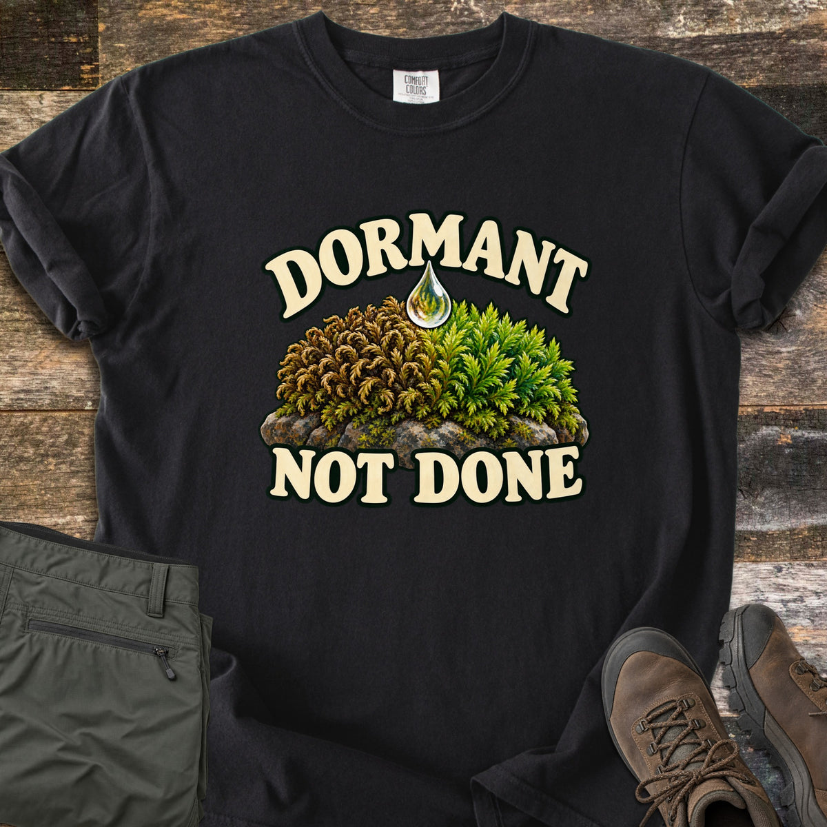 Dormant Not Done Comfort Colors T-shirt in Black, laid flat. Cream letters read Dormant over moss on a rock, dry brown on one side and green on the other, with a water drop above and Not Done underneath. Folded olive pants and brown hiking boots lie on rustic wood.