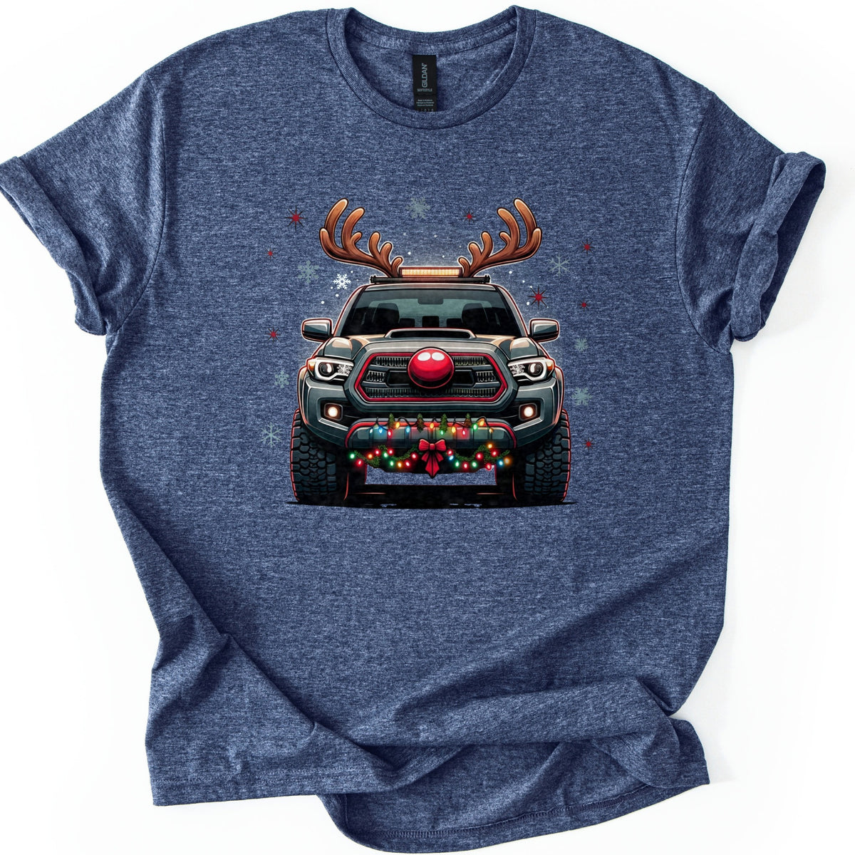 Santa Truck Comfort Colors T-shirt in Heather Navy, laid flat. A front-facing dark gray pickup truck with reindeer antlers, a round red nose on its grille, and colored lights with a red bow on the bumper, amid snowflakes and red stars. No words. Sleeves cuffed, on white.