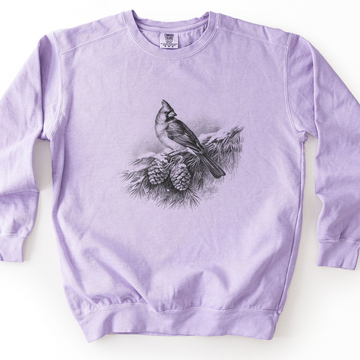 Cardinal On A Snowy Bough Comfort Colors crewneck sweatshirt in Orchid, laid flat. The chest shows a grayscale sketch of a cardinal perched on a pine bough dusted with snow, two pinecones hanging beneath and snowflakes around it, with no words. It lies flat on a white surface.