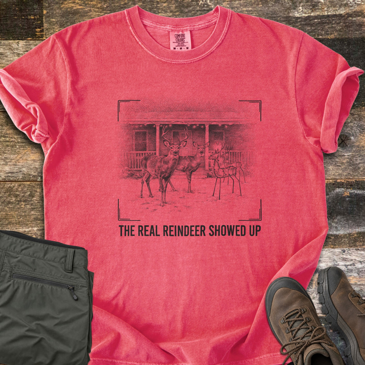 The Real Reindeer Showed Up Comfort Colors T-shirt in Watermelon, laid flat. A sketch in camera viewfinder corners shows a buck and doe in snow by a wire reindeer decoration at a porch, above the words The Real Reindeer Showed Up. Sleeves are rolled; olive pants and brown boots lie on weathered wood.
