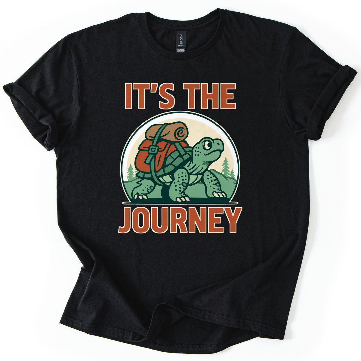 Slow Turtle Hiker Journey Comfort Colors T-shirt in Black, laid flat. A green cartoon turtle carrying a red backpack with a rolled sleeping bag walks past pine trees, framed by the words It's the Journey in rust-orange letters. Sleeves are cuffed on a white background.