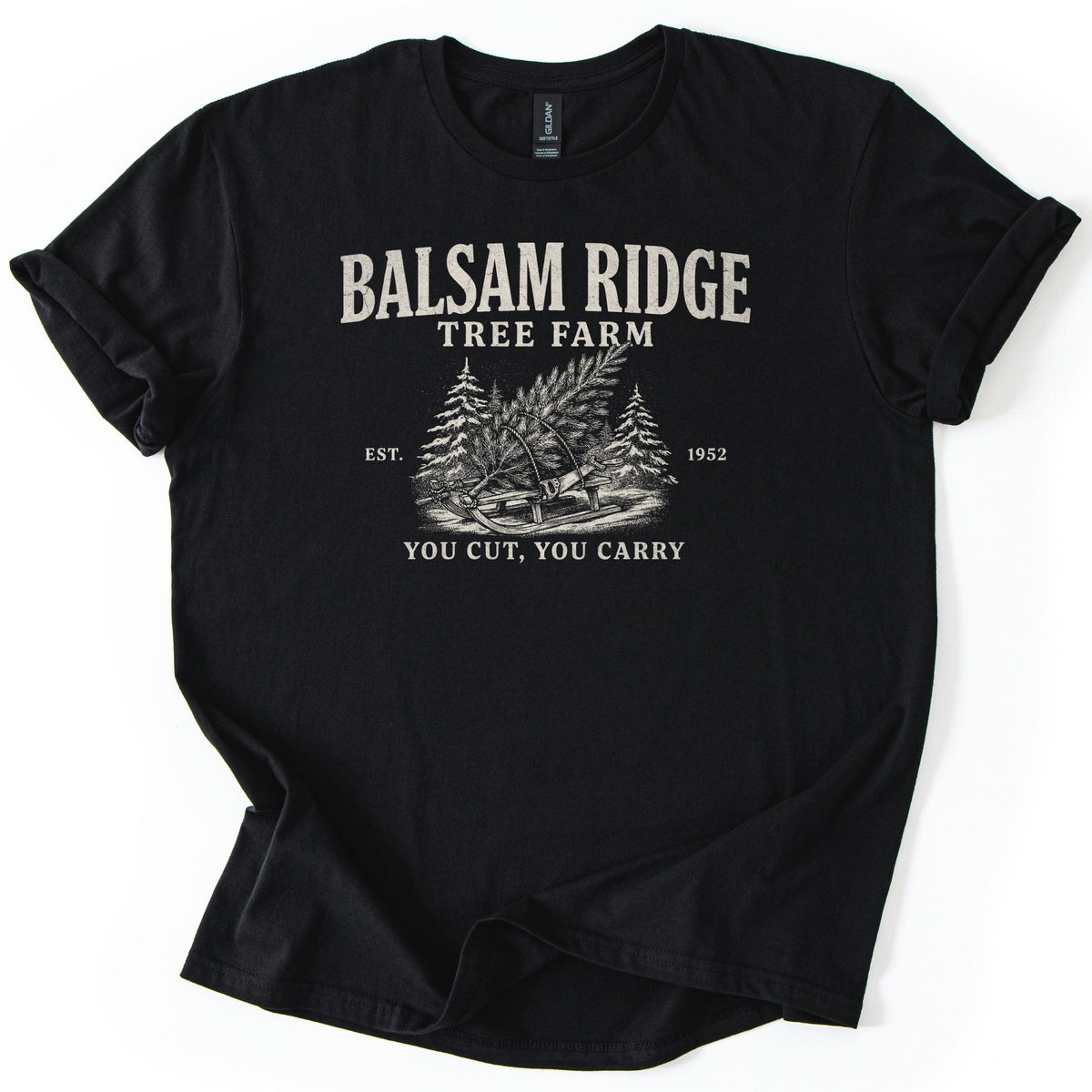 Balsam Ridge Tree Farm Comfort Colors T-shirt in Black, laid flat. Cream print reads Balsam Ridge Tree Farm above an engraved-style fir tree tied to a wooden sled between snowy pines, flanked by Est. 1952, with You Cut, You Carry below. Sleeves cuffed on a white surface.