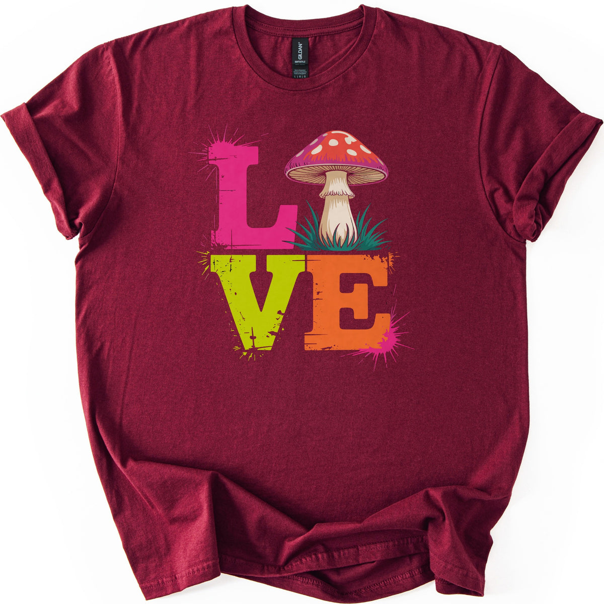 Mushroom Love Comfort Colors T-shirt in Maroon, laid flat. Distressed block letters spell LOVE, with a pink L, a red white-spotted mushroom in tufts of grass as the O, a lime green V and an orange E, and pink paint splatters. Sleeves are cuffed on a white background.
