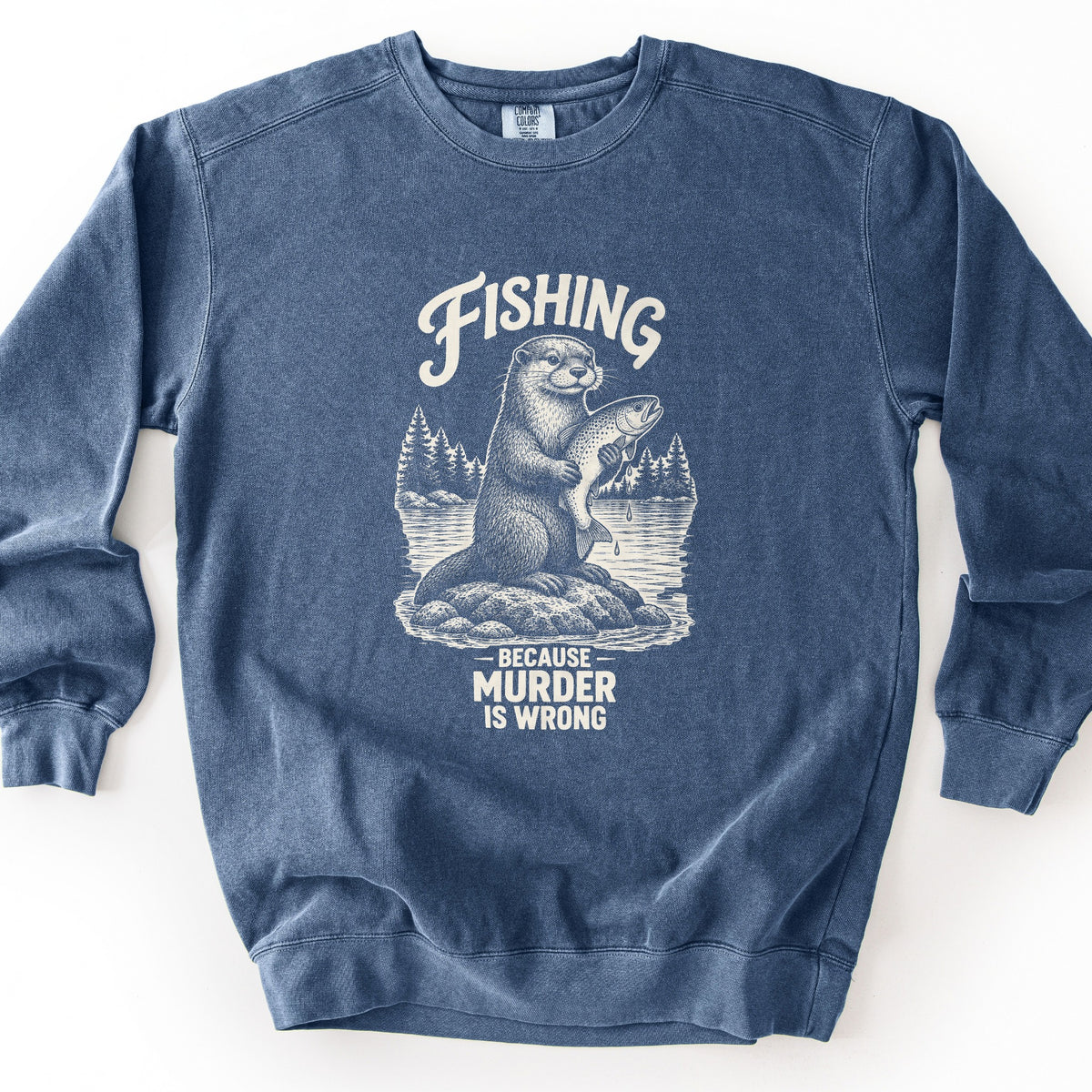 Fishing Because Murder Is Wrong Comfort Colors crewneck sweatshirt in Denim, laid flat. A cream drawing of an otter on a rock in a lake holds up a dripping trout, with pine trees behind it. Fishing is printed above it and Because Murder Is Wrong below. Set on a white surface.