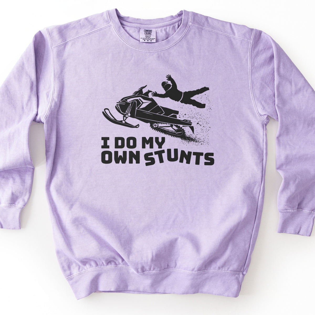 I Do My Own Stunts Comfort Colors crewneck sweatshirt in Orchid, laid flat. A black print shows a helmeted rider launched from an airborne snowmobile with a burst of snow, above the words I DO MY OWN STUNTS. The sweatshirt lies on a white surface.