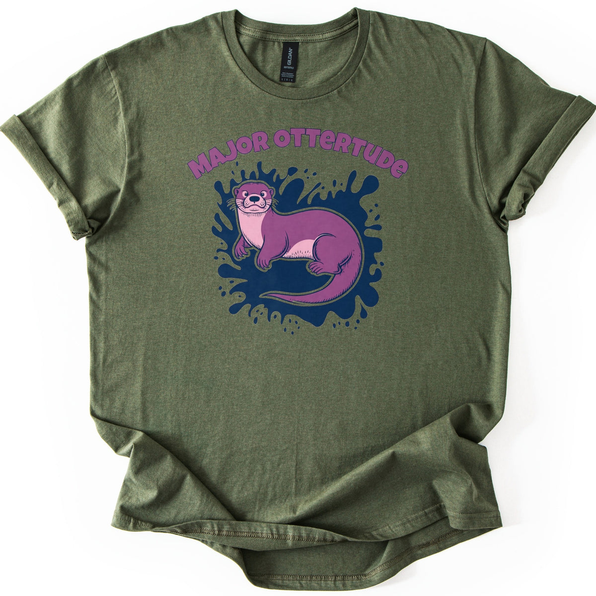 Major Ottertude Comfort Colors T-shirt in Military Green, laid flat. A purple cartoon otter with a pink chest lies on a navy blue paint splash below arched purple letters reading MAJOR OTTERTUDE. The shirt has cuffed sleeves and lies on a white background.