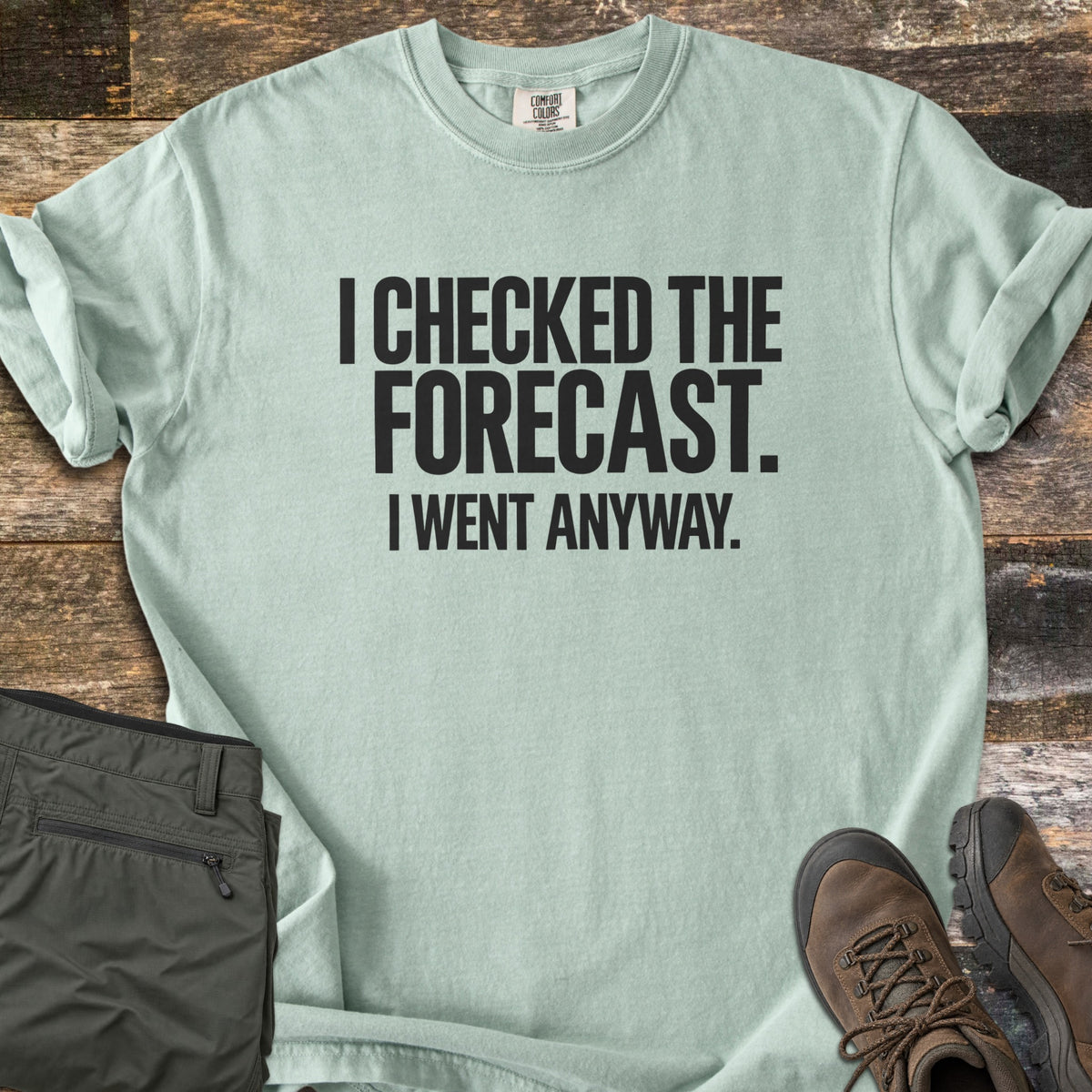 Checked The Forecast Comfort Colors T-shirt in Bay, laid flat. Black block letters read I CHECKED THE FORECAST. I WENT ANYWAY. The sleeves are cuffed, with folded olive hiking pants and brown hiking boots beside it on weathered wood planks.