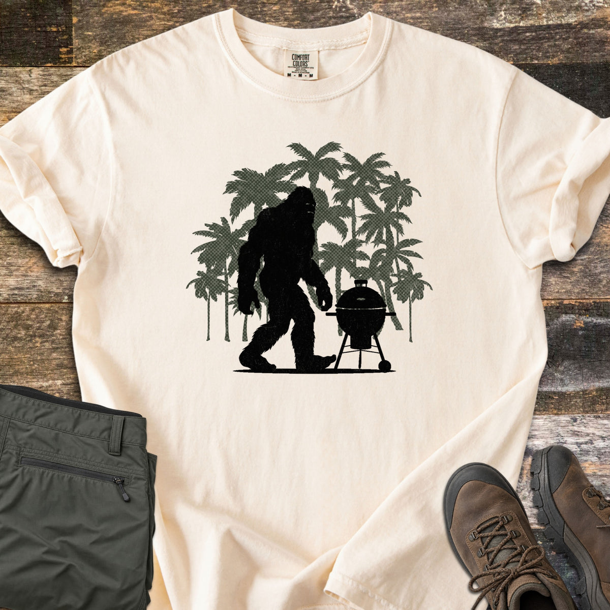 Squatch BBQ Smoker Comfort Colors T-shirt in Ivory, laid flat. A black Bigfoot silhouette walks past a kettle grill on wheels, set against dark green palm tree silhouettes; no words are printed. The shirt lies on weathered wood beside olive pants and brown hiking boots.