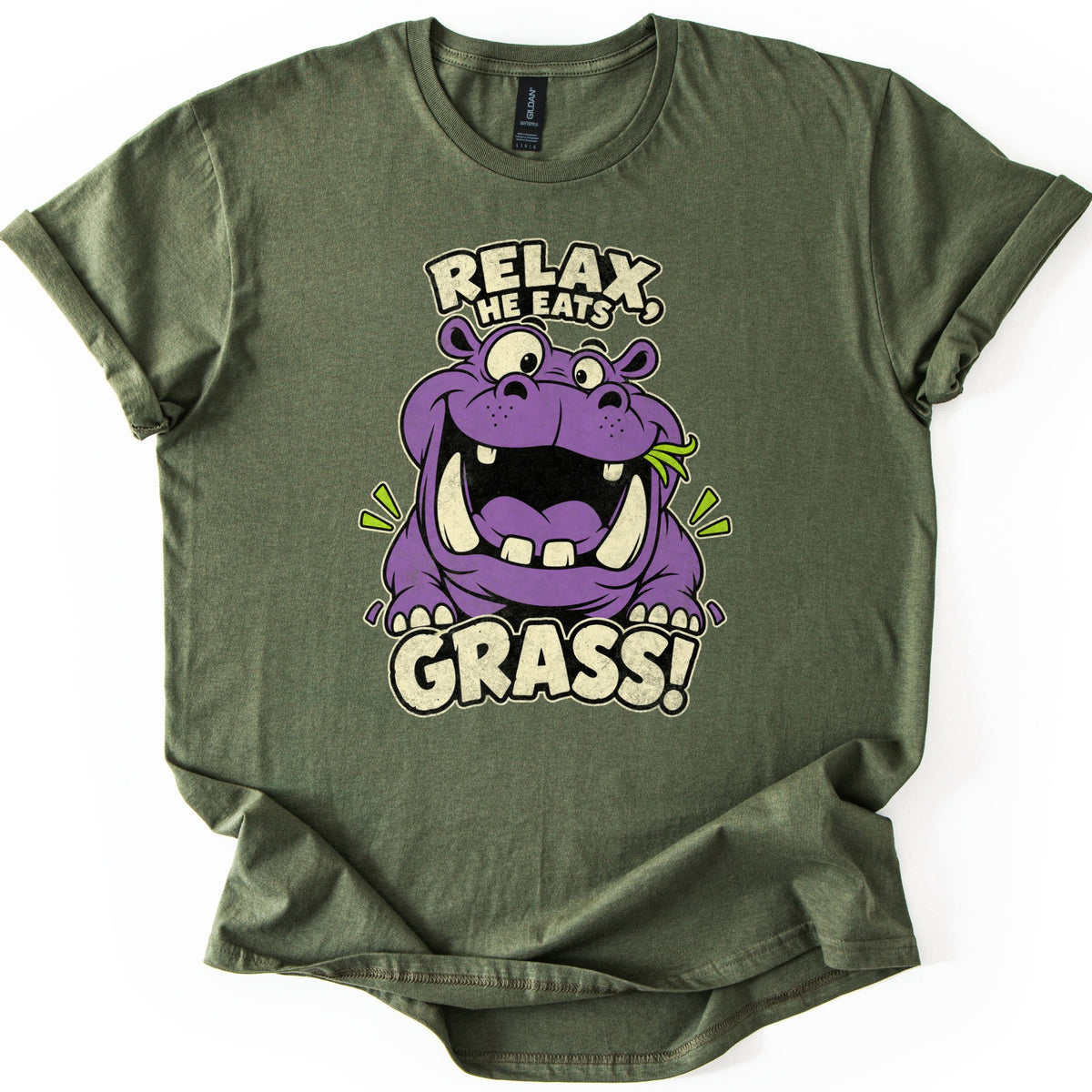 Relax He Eats Grass Comfort Colors T-shirt in Military Green, laid flat. A grinning purple cartoon hippo with big tusks and a blade of grass in its mouth sits between the words RELAX, HE EATS and GRASS! in cream letters. The shirt lies on white with rolled sleeves.