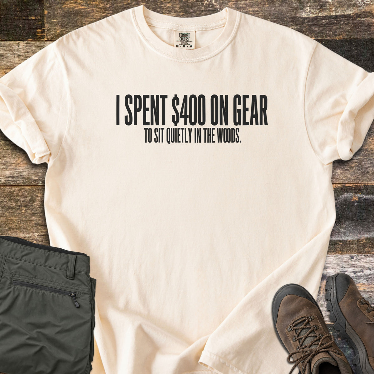 Gear To Sit Quietly Comfort Colors T-shirt in Ivory, laid flat. Black text reads I SPENT $400 ON GEAR, with TO SIT QUIETLY IN THE WOODS. below. Sleeves are rolled on a rustic wood surface, beside folded olive hiking pants and brown hiking boots.