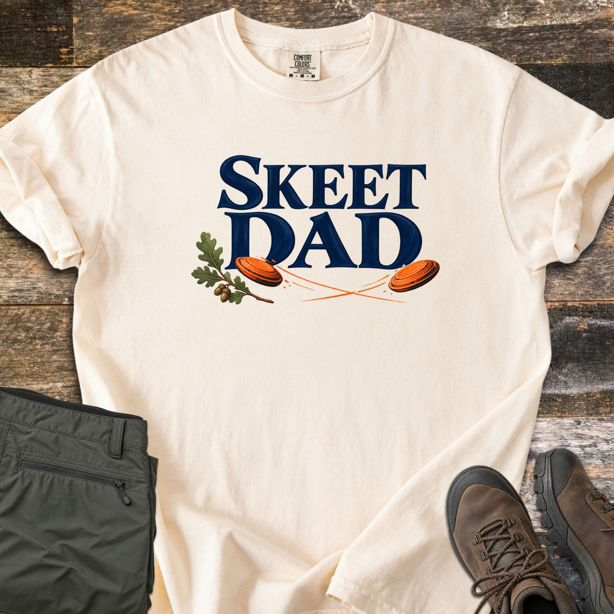 Skeet Dad Comfort Colors T-shirt in Ivory, laid flat. Navy text reads Skeet Dad above two orange clay targets with motion trails and an oak sprig with acorns. Sleeves are rolled on a wood floor beside folded olive cargo pants and brown hiking boots.