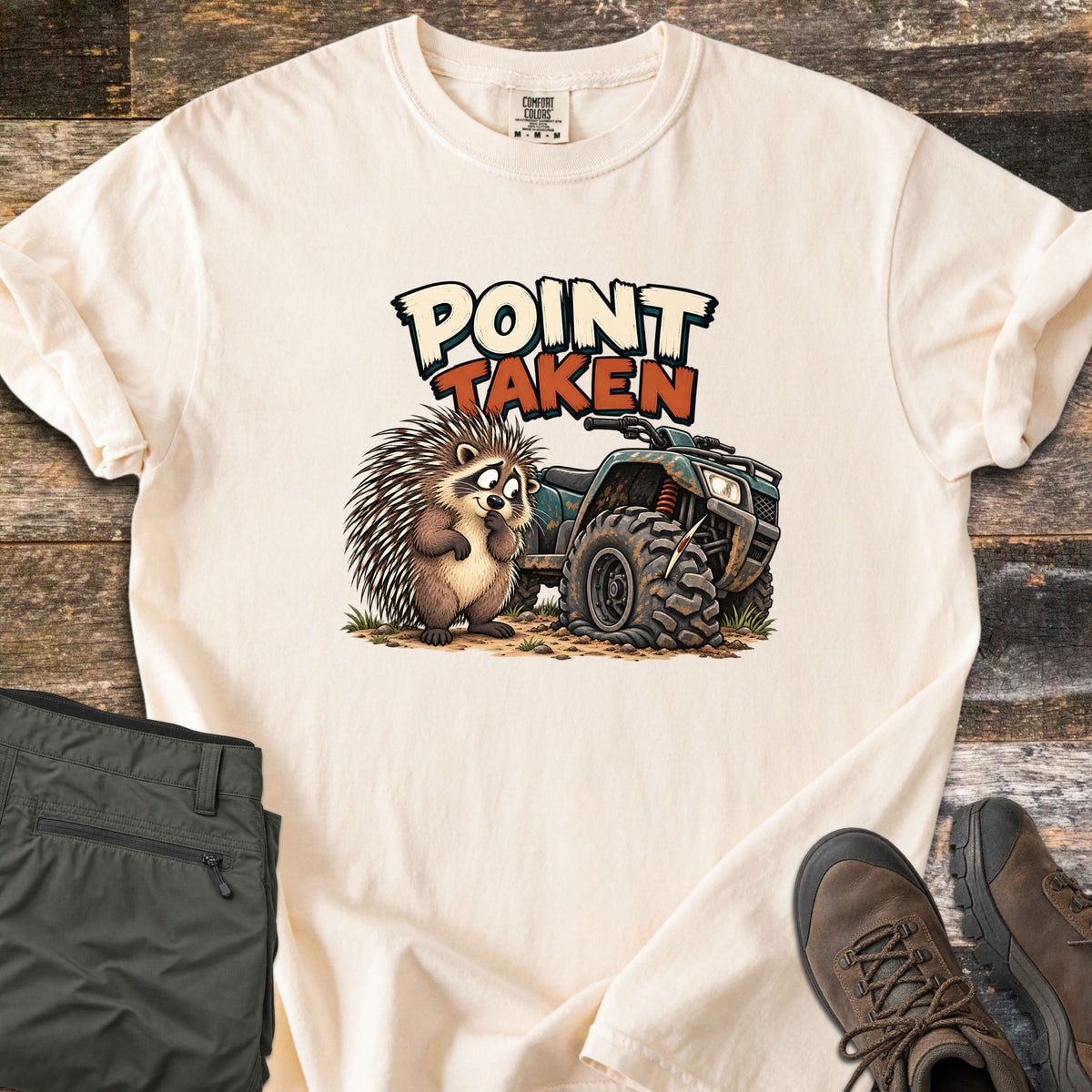 Point Taken Comfort Colors T-shirt in Ivory, laid flat. A cartoon porcupine looks sheepishly at a muddy ATV with a flat front tire pierced by a quill, under the words Point Taken. Folded olive pants and brown hiking boots sit beside it on weathered wood.