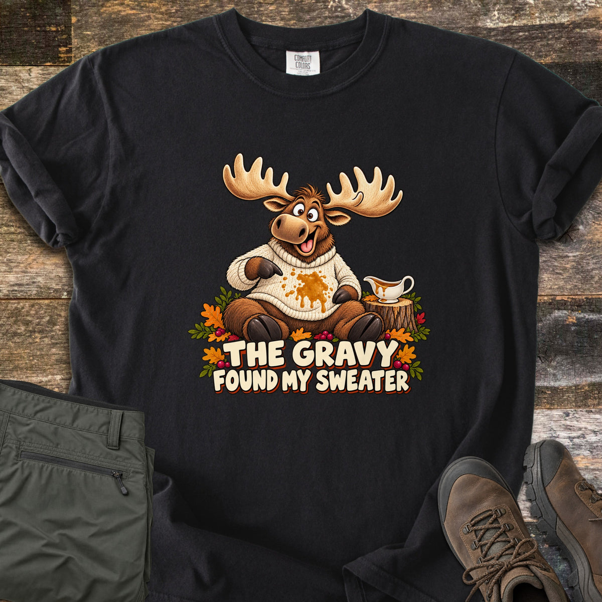The Gravy Found My Sweater Comfort Colors T-shirt in Black, laid flat. A cartoon moose in a cream knit sweater points at a gravy stain beside a gravy boat on a stump, above the words The Gravy Found My Sweater. Olive pants and brown boots sit on a wood floor.