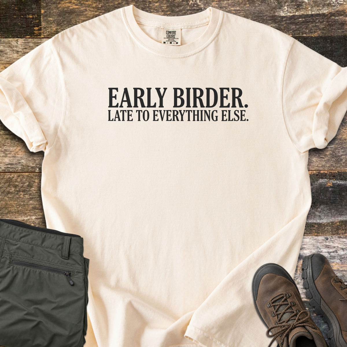 Early Birder Comfort Colors T-shirt in Ivory, laid flat. Black serif text across the chest reads EARLY BIRDER. LATE TO EVERYTHING ELSE. The shirt has rolled sleeves and lies on weathered wood planks beside folded olive hiking pants and brown hiking boots.