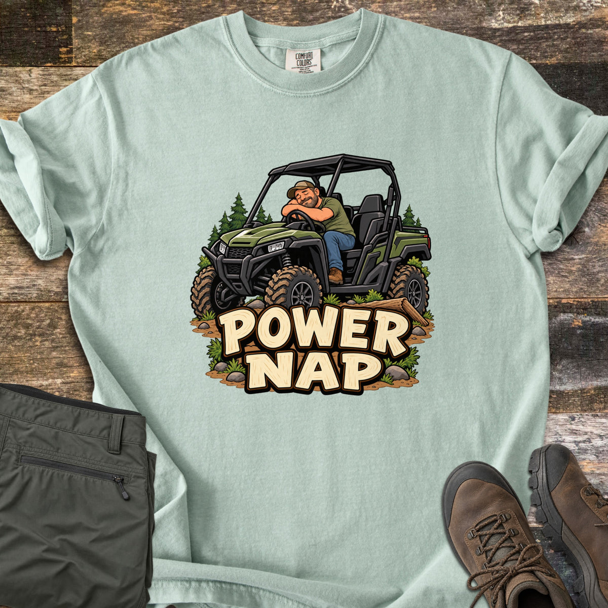 Power Nap Comfort Colors T-shirt in Bay, laid flat. A cartoon man in a cap naps with his head on the steering wheel of a green side-by-side UTV among pine trees, above the words Power Nap. Folded olive pants and brown hiking boots sit on a weathered wood surface.